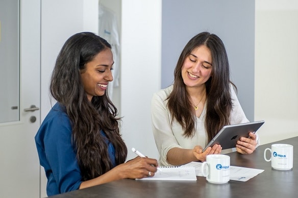 Two female employees are exchanging ideas at a table during a meeting. The one on the right is holding an iPad and the one on the left is writing on a notepad.